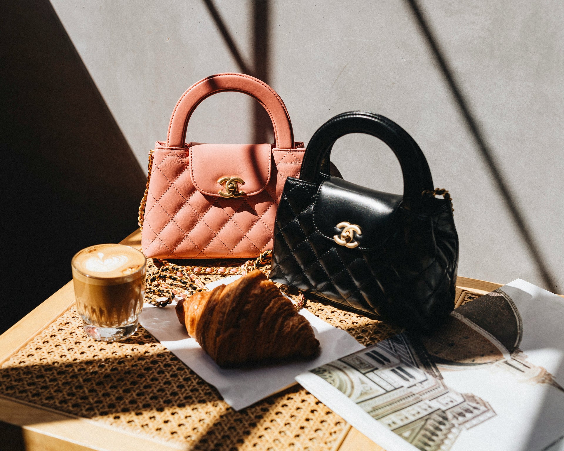 Two handbags, one pink and one black, on a table with a coffee and croissant.